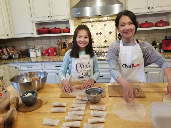 A woman and her daughter in a kitchen, wearing aprons and making food